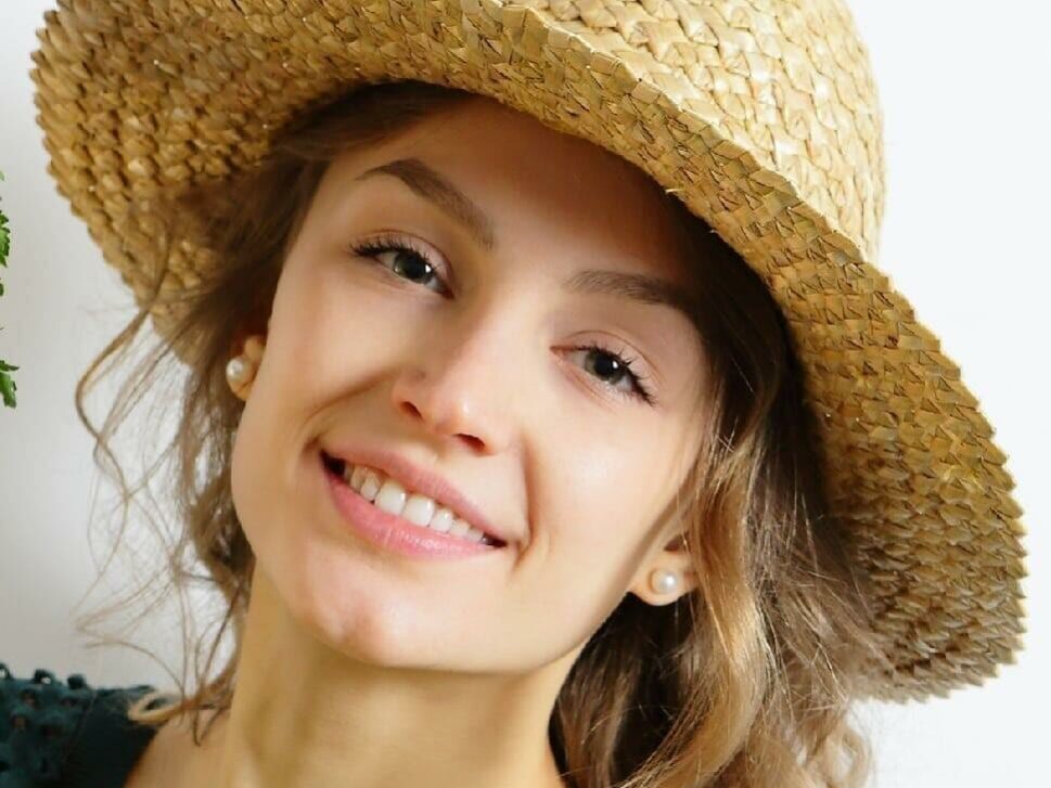 sunscreenblog image-100 A woman with long, wavy hair smiles at the camera. She is wearing a wide-brimmed straw hat and small pearl earrings. The background is plain white, highlighting her cheerful expression and casual, summery appearance.