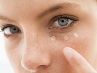 Foundation blog A close-up of a person with blue eyes applying three dots of light-colored concealer under their left eye using their finger. The skin appears natural with freckles, and the background is out of focus.