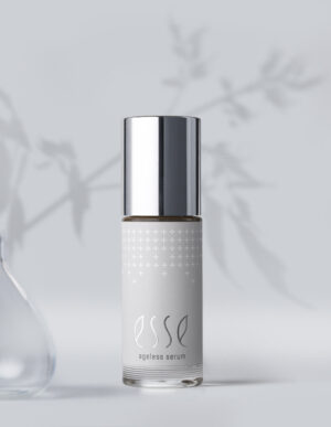 A silver-capped bottle of Esse Ageless Serum stands on a light surface, with a blurred shadow of leaves in the background and part of a glass vase visible on the left.