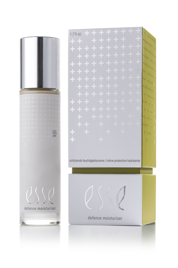 A bottle of Esse Defence Moisturiser with a silver cap stands beside its matching silver and yellow box, both displaying a minimalist design with crosses and the product name: Esse Defence Moisturiser.
