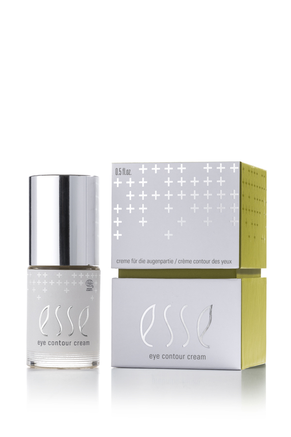 A small bottle of Esse Eye Contour Cream with a silver cap sits beside its white and green box, decorated with minimalist plus signs and product details in English, French, and German.