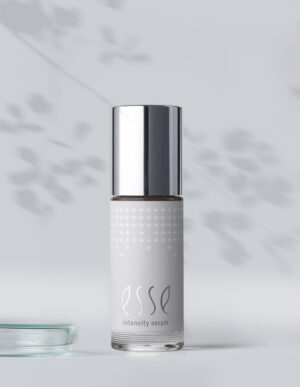 A bottle of Esse Intensity Serum with a metallic cap stands on a light surface, soft leaf shadows in the background, and the edge of a glass dish partially visible on the left.