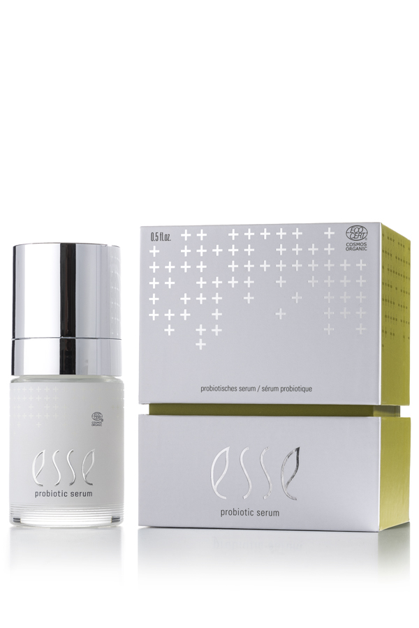A bottle of Esse Probiotic Serum with a shiny silver cap sits beside its white-and-green box, which features silver cross patterns along with the product name and details.