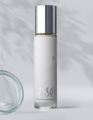 A tall, sleek bottle with a silver cap labeled "Esse Restorative Moisturiser" stands upright on a pale surface, with a blurred glass container and soft shadows in the background.