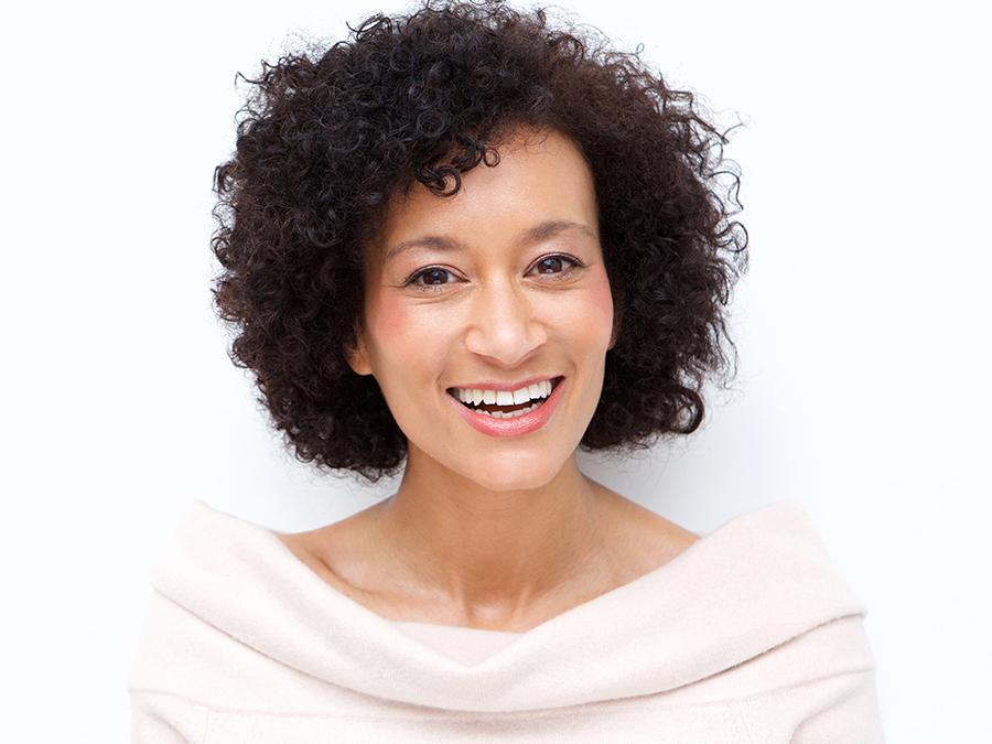 A woman with short curly hair smiles warmly at the camera. She is wearing an off-the-shoulder, light-colored top. The background is light and uncluttered, drawing attention to her joyful expression.