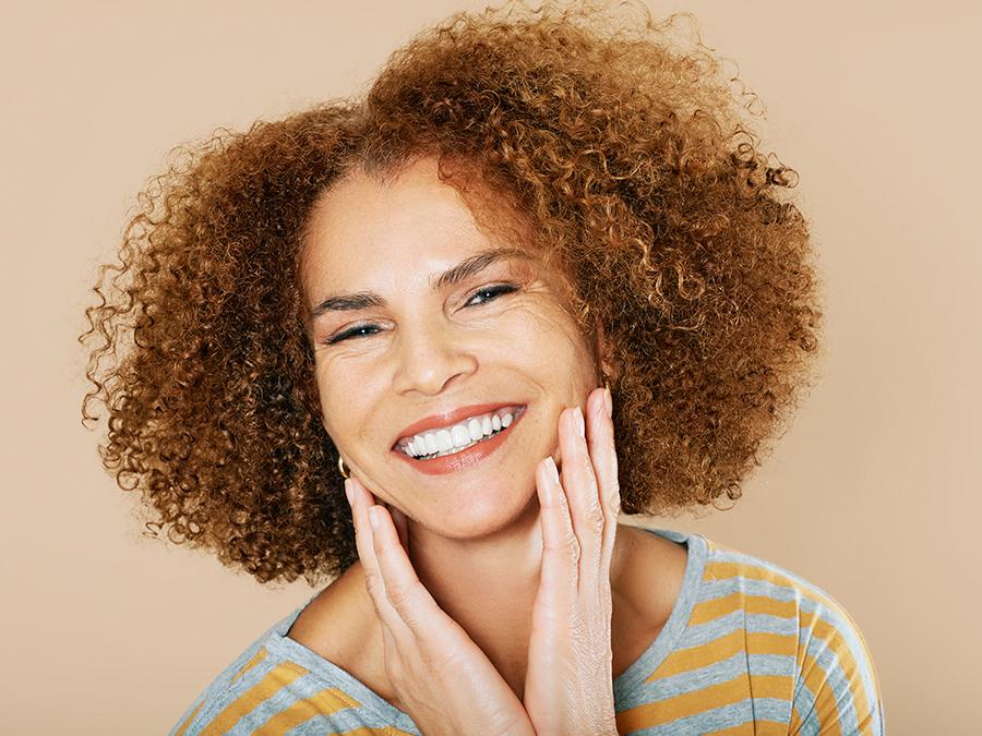 A smiling woman with curly hair touches her face with both hands. She is wearing a gray and yellow striped shirt, and stands against a beige background.