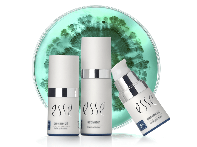 Aesthetic_blog_image-01 Three Esse skincare product bottles are displayed in front of a dish with a green and teal gradient pattern. The products include pre-serum oil, activator, and a third bottle with a pump dispenser. All bottles are white with silver accents and branded "Esse.