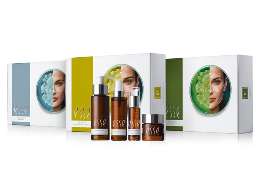 3_TRIAL_PACKS-825x620px A display of skincare products from the brand Esse, featuring various bottles and containers in front of three boxes with images of a face on each. The packaging colors are predominantly white, blue, yellow, and green.