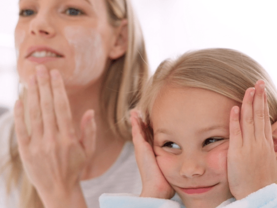 Screenshot_2023-11-14_100501 A woman is applying skincare cream to her face while smiling. Next to her, a young girl with long blonde hair is mimicking the action, touching her cheeks with a playful smile. Both are wearing casual, light-colored clothing, and the background is softly blurred.