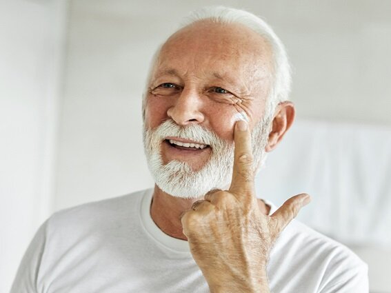 An elderly man with white hair and a beard smiles as he applies a cream to his cheek using his finger. He is wearing a white shirt and appears to be in a well-lit room, possibly a bathroom or bedroom.