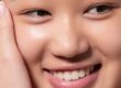 Close-up of a smiling person with clear, smooth skin, natural makeup, and white teeth, gently touching their cheek with one hand.