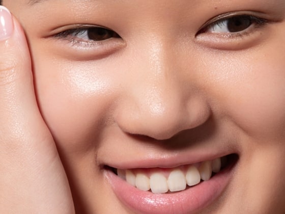 Close-up of a smiling person with clear, smooth skin, natural makeup, and white teeth, gently touching their cheek with one hand.