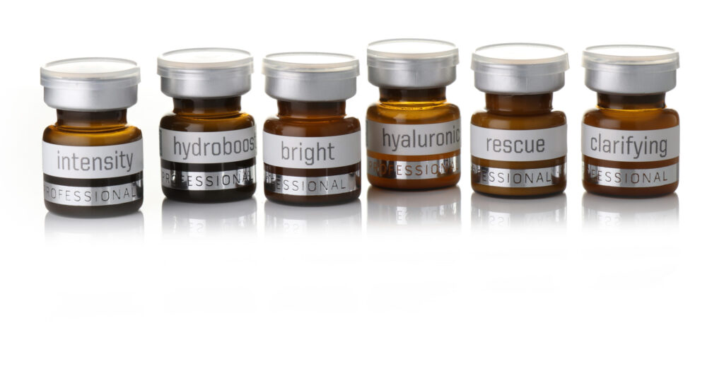 Six small amber glass jars with silver lids, each labeled with a different skincare function: intensity, hydroboost, bright, hyaluronic, rescue, and clarifying, arranged in a row on a white background.
