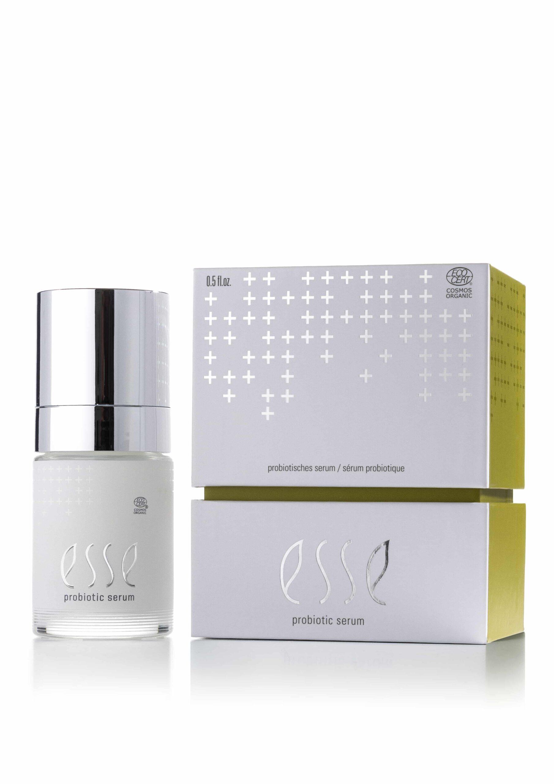 A bottle of Esse Probiotic Serum with a shiny silver cap stands beside its white box, featuring metallic silver cross designs and gold sides. The label displays the product name, “Esse Probiotic Serum.”.