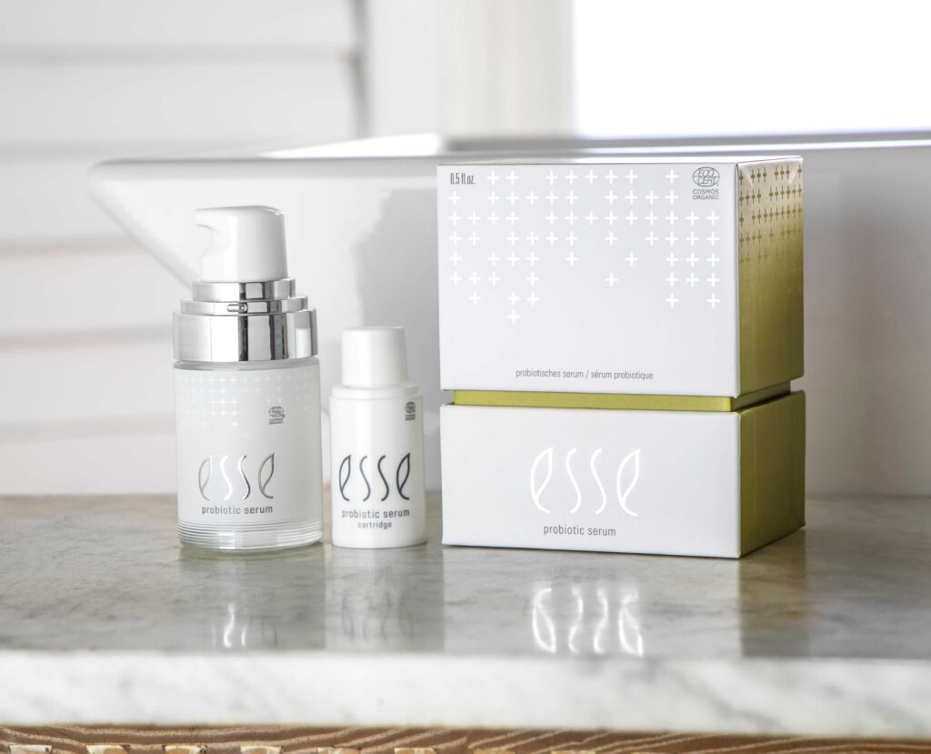 Two bottles of Esse Probiotic Serum and a matching white and gold box are displayed on a marble countertop, with a window and white wall in the background.