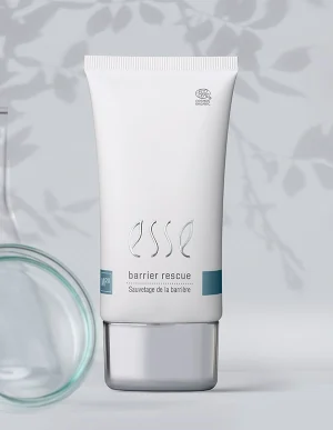 A white tube labeled “Import placeholder for 57617” stands upright on a reflective surface, with a blurred glass container and leafy shadows in the background. The packaging features minimalist text and a silver cap.